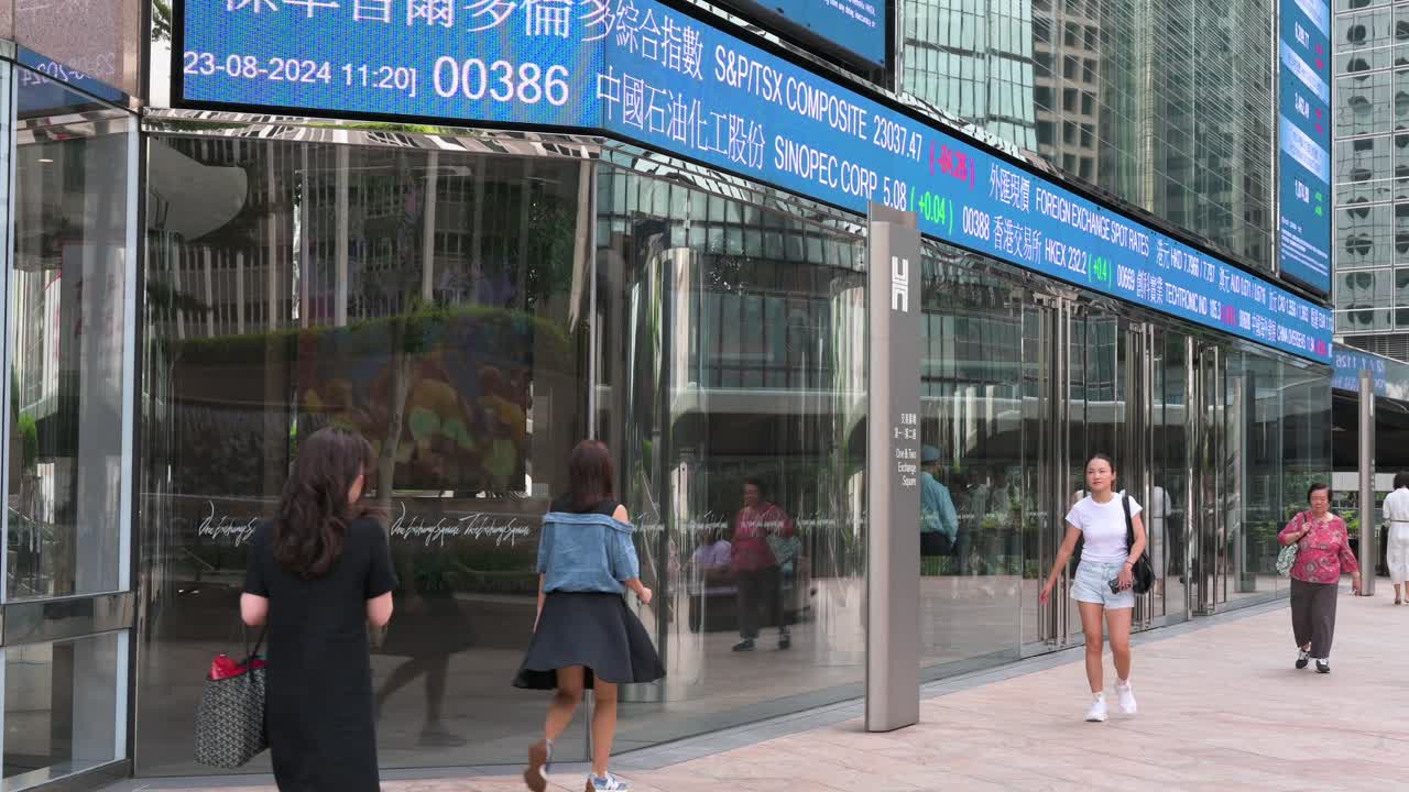 In Hong Kong's financial district, pedestrians walk past a moving screen at Exchange Square, showcasing negative stock ticker symbols at the home of the Hong Kong Stock Exchange (HKEX).