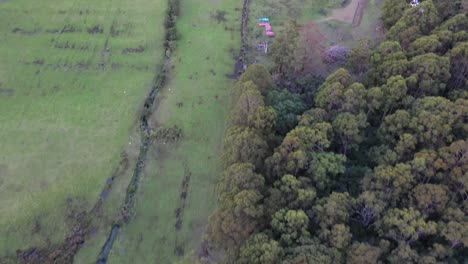 Slow-aerial-drone-pan-over-grassy-plains-and-forest,-slow-setting-sunset-of-island-beach-at-dusk,-purple-and-orange-sky-and-mountain-range-with-shacks-and-clouds