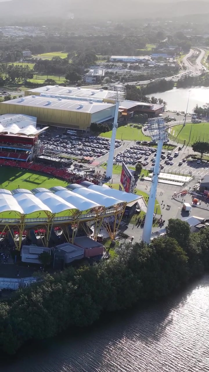 High-angle drone shot reveals a large sports arena beside a shimmering river under bright sunlight.