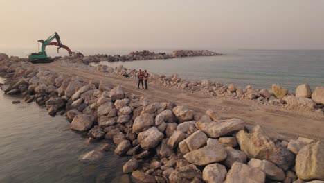 two workers stand on a new breakwater and two excavator machines in the background during sunset