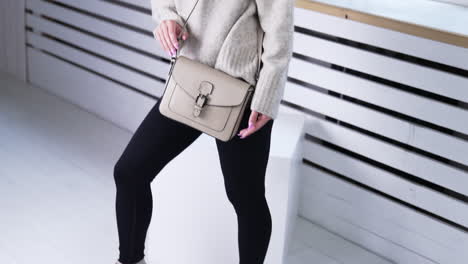 model in warm sweater and black pants holding a grey bag. female demonstrating fashionable shoes indoors. white backdrop.