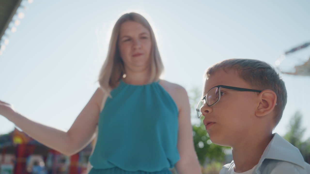 White mother and son in park during weekend outing under bright sunlight, mother in teal top stands behind son with glasses near carnival rides and palm trees, candid serious expressions, protective