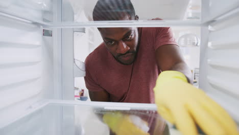 view looking out from inside empty refrigerator as man wearing rubber gloves cleans shelves