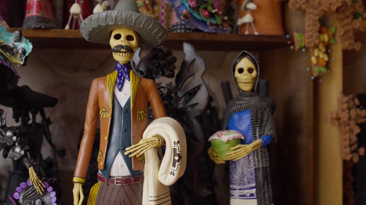 Fargerike håndlagde Catrina-skulpturer utstilt på en hylle i et håndverksverksted, malt med vakre, subtile farger.