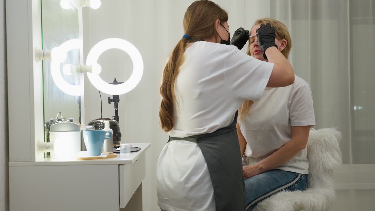 Beautician wearing gloves arranges blond client eyebrow under bright ring light in modern salon, with various grooming tools and blue mug visible on table, showcasing clean and professional workspace