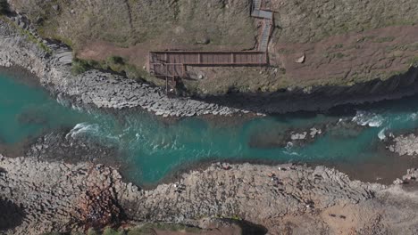 Blue-glacial-river-in-Stuðlagil-canyon-with-tourist-viewing-platform-on-cliff,-aerial
