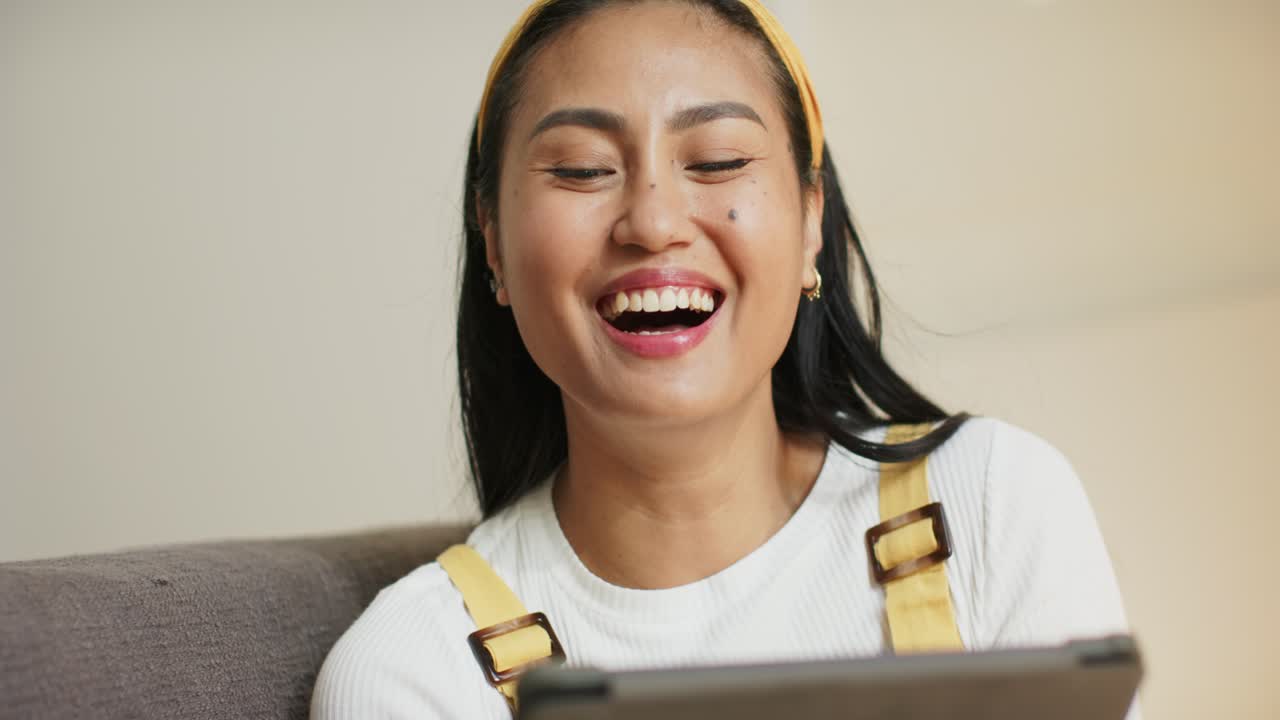 リビングのグレーのソファでタブレット画面をタップし、コンテンツをスワイプし、笑っている中年アジア人女性