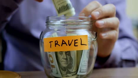 hombre caucásico poniendo un frasco de vidrio puesto en una mesa de cajón con sus ahorros para viajes futuros