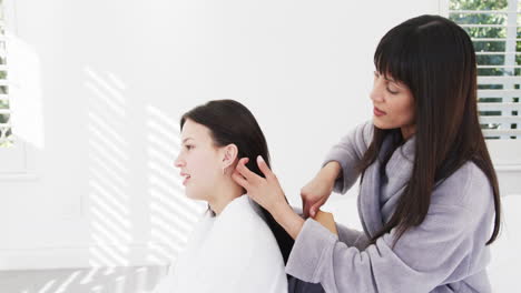 Happy-biracial-mother-in-bathrobe-brushing-hair-of-smiling-adult-daughter-in-sunny-room,-slow-motion