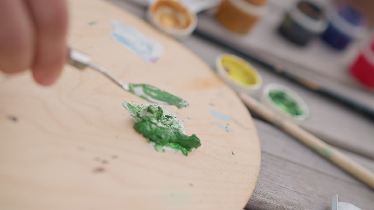 Palette con pigmento verde vivido miscelato su tavola di legno con coltello e spatola, barattoli di colore e pennelli sullo sfondo, allestimento da studio all'aperto per murales su strada, artista che miscela acrilico denso per creare texture