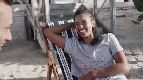 Happy-diverse-couple-sitting-in-deckchairs-talking-outside-beach-house-in-the-sun,-in-slow-motion