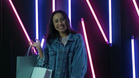 young girl in blue denim jacket with shopping bags on neon lamps background.