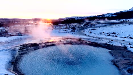 beautiful panoramic view of a geyser in geysir, iceland. smoke is getting out of the geyser with people walking all around the geyser, under the red and orange sunset.