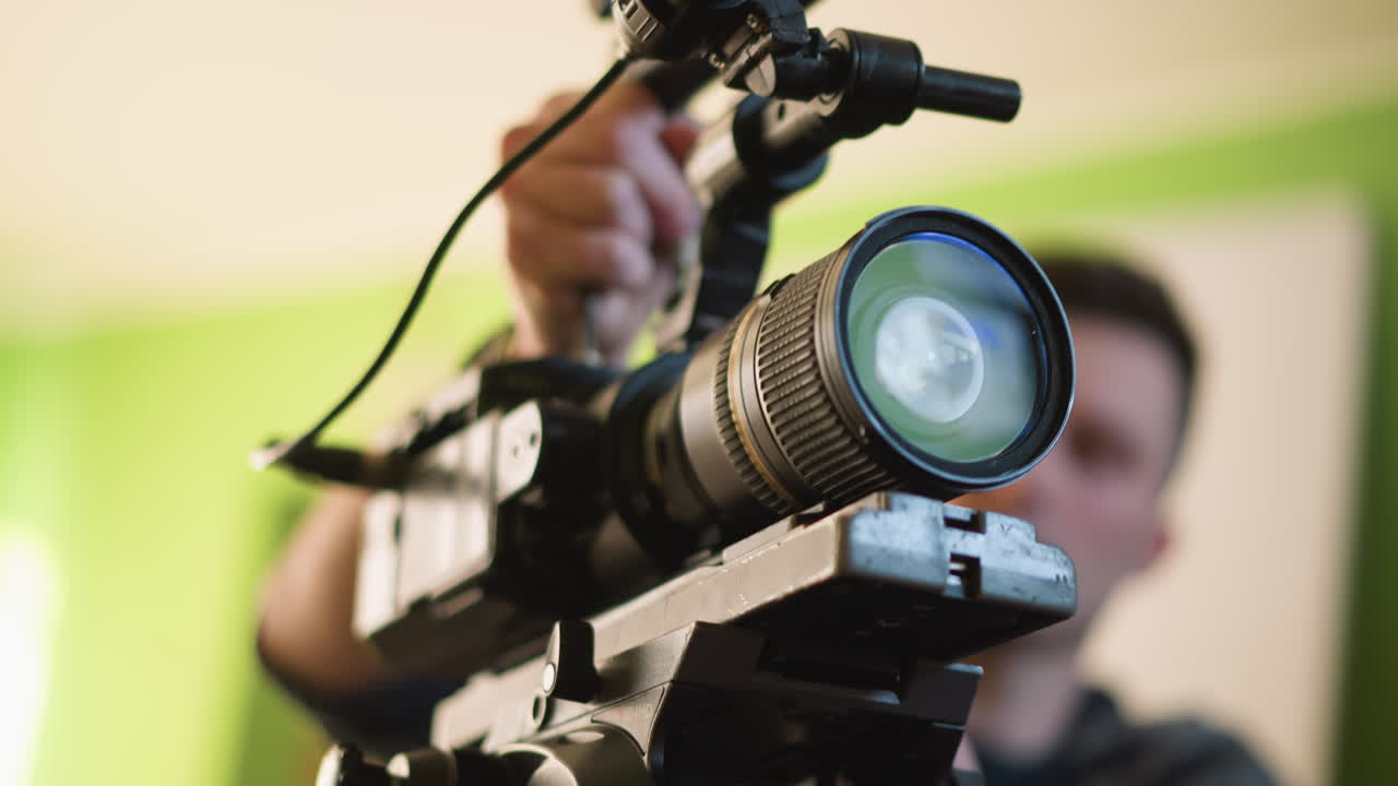 Close-up of camera tripod plate, cameraman adjusting camera in background, blurry green environment, focus on tripod components, lens, and microphone