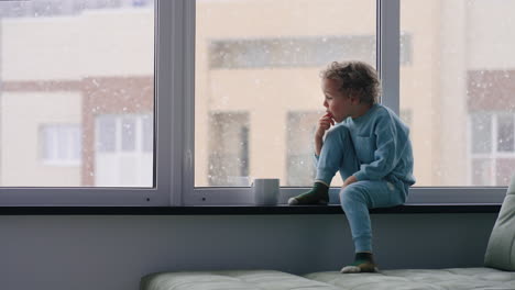 Fröhlicher-Kleiner-Junge-Probiert-Heiße-Schokolade-Auf-Der-Fensterbank-Zu-Hause,-An-Einem-Wintertag-Fällt-Schnee-Auf-Die-Straße