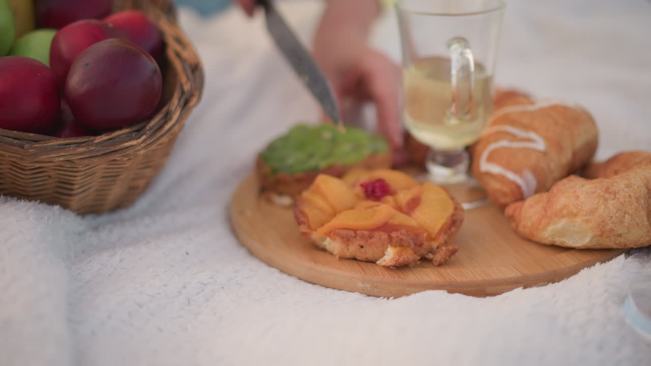 tavola da picnic illuminata dal sole con croissant e torta, bicchiere di succo accanto a un cesto di vimini pieno di prugne, mani adulte che tagliano una crostata di frutta su un tagliere di legno, coperta morbida, luce mattutina, atmosfera da brunch accogliente