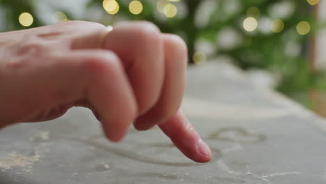 Hand view drawing love shape on dusty box using wet finger with blurred glowing Christmas tree lights in background, capturing heartfelt creative moment during cozy festive indoor holiday celebration