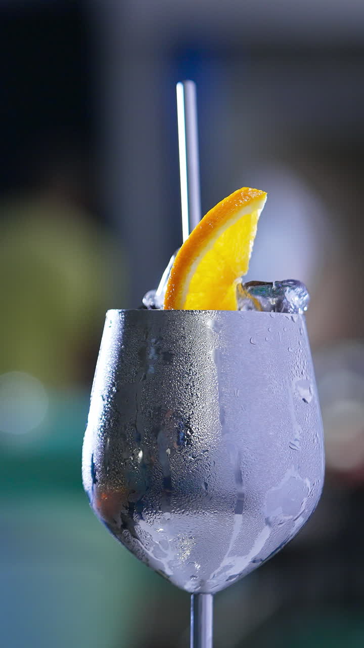Rotating metal wineglass with misted surface. Cocktail with ice and orange slice on top and metal straw inside. Close up. Blurred backdrop. Vertical video