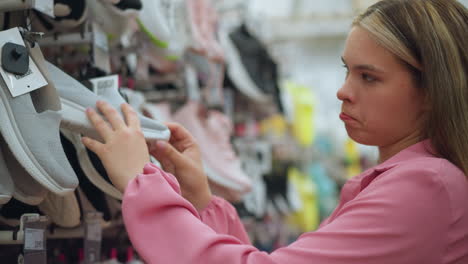 lady in pink dress in shoe store examines an ash sneaker, focusing closely on the sole with a facial expression of uncertainty, the shelf displays various styles of shoes, slightly blurred