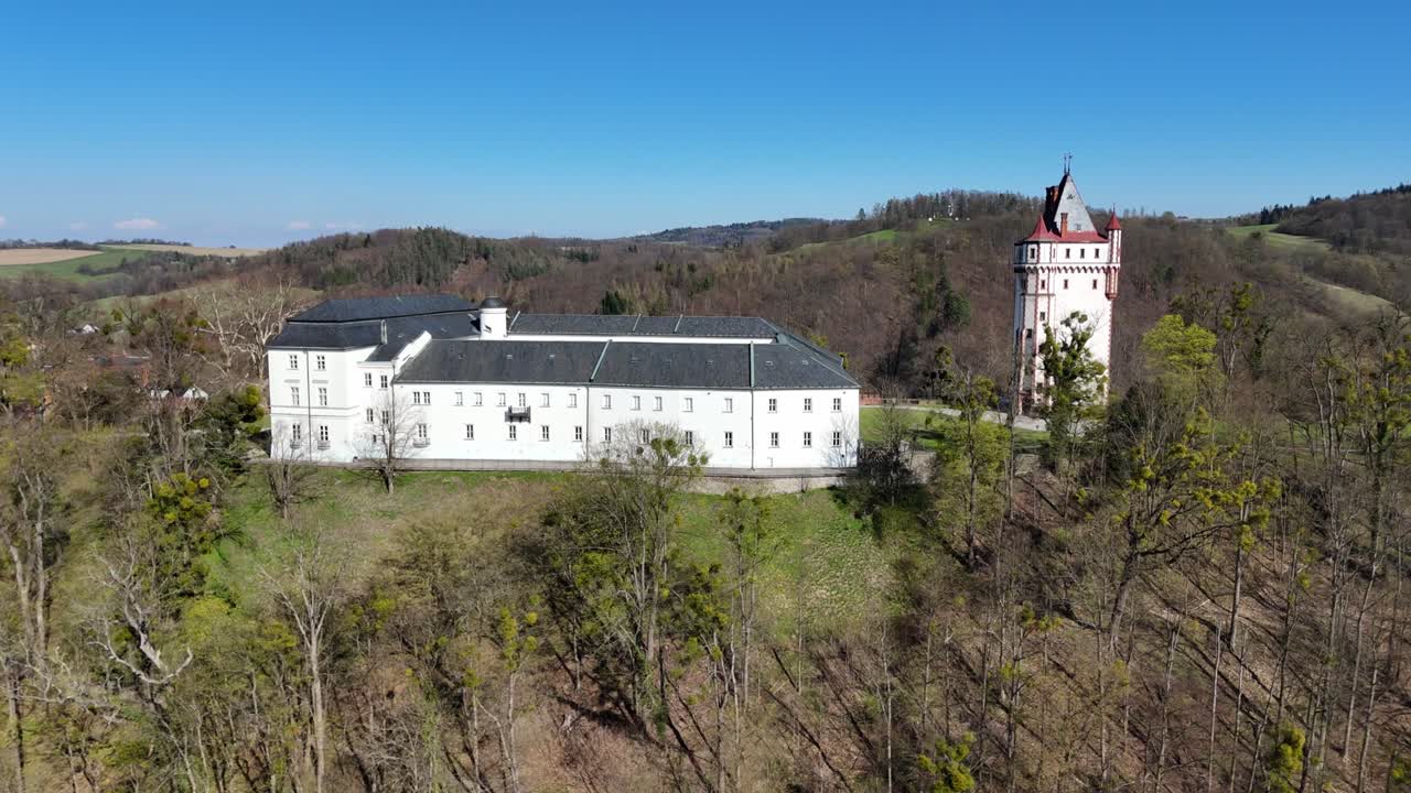 Boční pohyb, zatímco dron pomalu klesá, odhaluje zámek Hradec nad Moravicí s bílou budovou a věží nad stromy a zvlněnou krajinou pod jasně modrou oblohou.