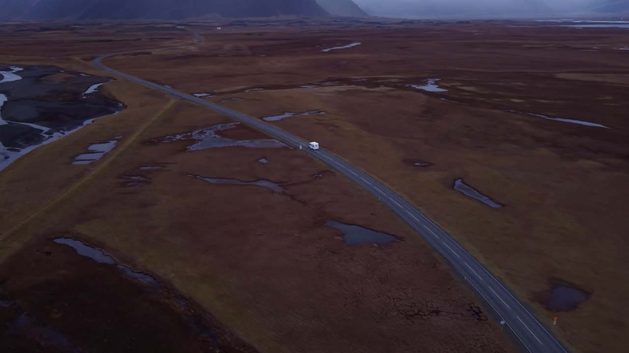 An expansive aerial shot captures a motorhome traveling along a road that cuts through a vast, rugged, and sparsely vegetated Icelandic landscape