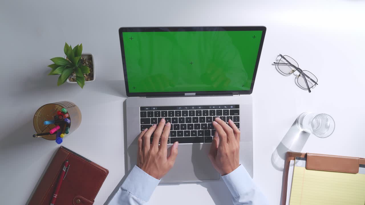 Top down view of a laptop computer with mock up green screen chromakey display on a wooden office desk next to notebook with pens, glasses, and a glass of water. Slow zoom out, close up