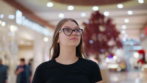joven con camiseta negra y gafas mueve la cabeza ligeramente, ajustando el cabello mientras se centra en algo en la distancia, el fondo presenta luces bokeh borrosas y un entorno vibrante de un centro comercial