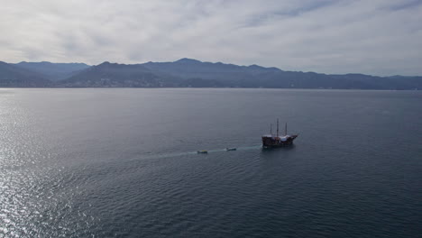 Drone-video-showing-a-pirate-ship-sailing-along-the-coasts-of-Puerto-Vallarta-and-Banderas-Bay,-orbiting-the-impressive-vessel,-showing-the-distant-shoreline-and-beautiful-backlit-sunlight