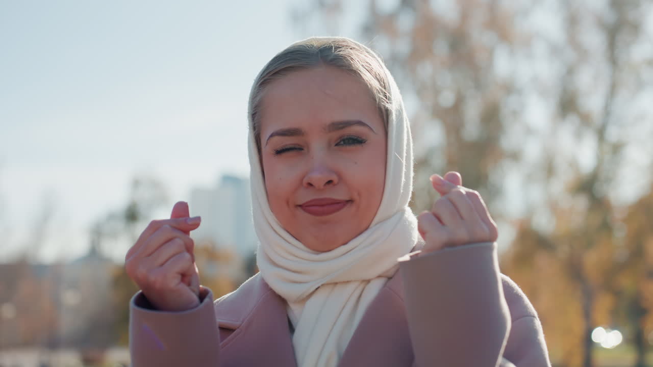Cuplikan influencer perkotaan. Wanita percaya diri membuat gestur mengundang. Pemandangan kota menangkap pose percaya diri bernuansa hangat. Influencer wanita memancarkan pesona dengan gestur hati jari dan latar belakang kota.