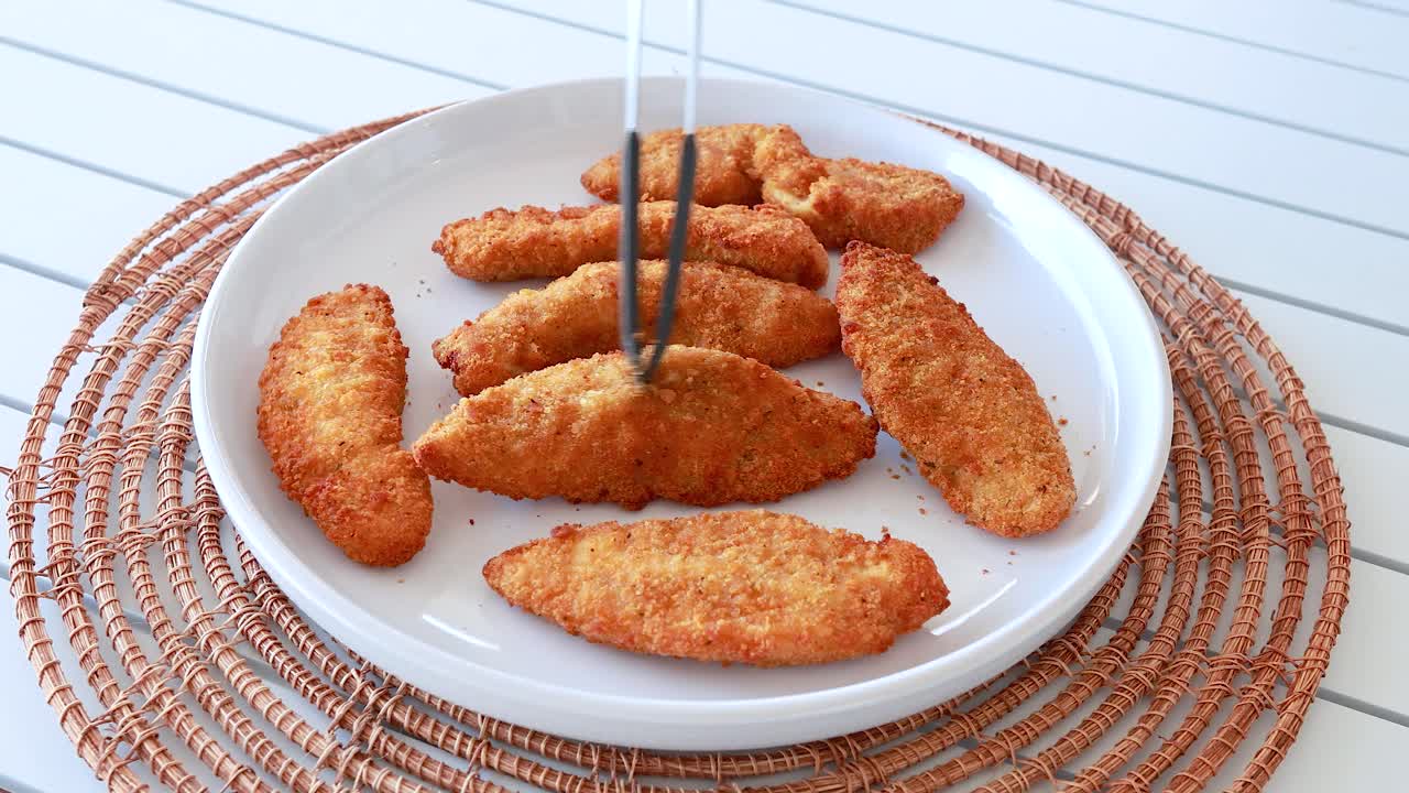 Tongs arrange golden fried chicken pieces on a white plate, set on a woven mat, under bright natural lighting