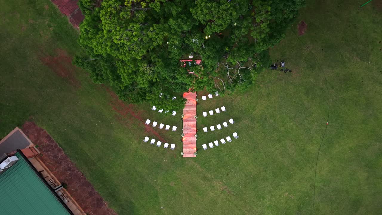 Volo discendente del drone dall'alto verso un allestimento per una cerimonia di matrimonio all'aperto con passerella in legno e sedie bianche.