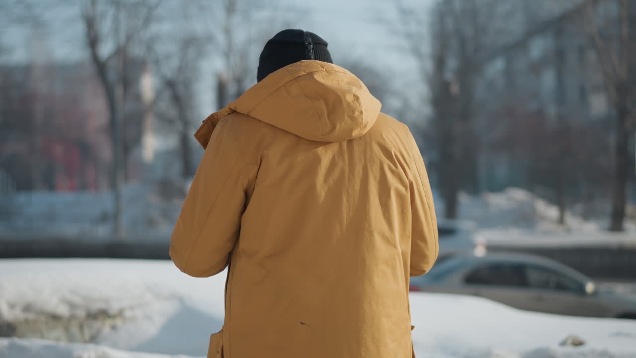 male artist walking on snow covered pedestrian path turns to frame shots with camera on neck while visible breath escapes under bright winter sun as busy street unfolds with passing cars