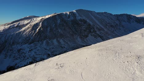 Ein-Einsamer-Mann-Mit-Schneeschuhen,-Der-An-Sonnigen-Tagen-In-Verschneiter-Winterberglandschaft-Bergauf-Geht,-Gipfel-Und-Klarer-Himmel-Im-Hintergrund,-Drohnenaufnahme