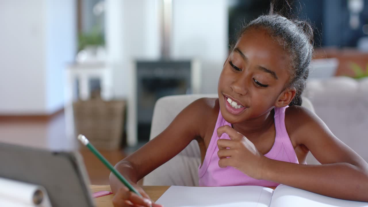 African American teen viewing tablet lesson to study, picking pencil writing in notebook at home