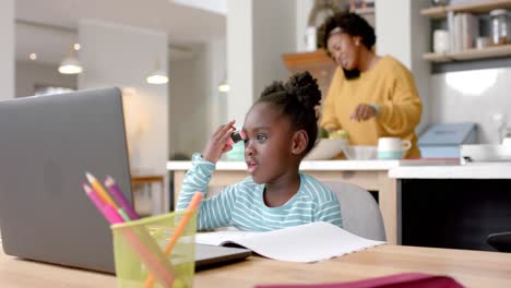 Niña-Afroamericana-Aprendiendo-En-Línea-En-La-Mesa-Usando-Una-Computadora-Portátil-Con-Su-Madre-En-La-Cocina,-Cámara-Lenta