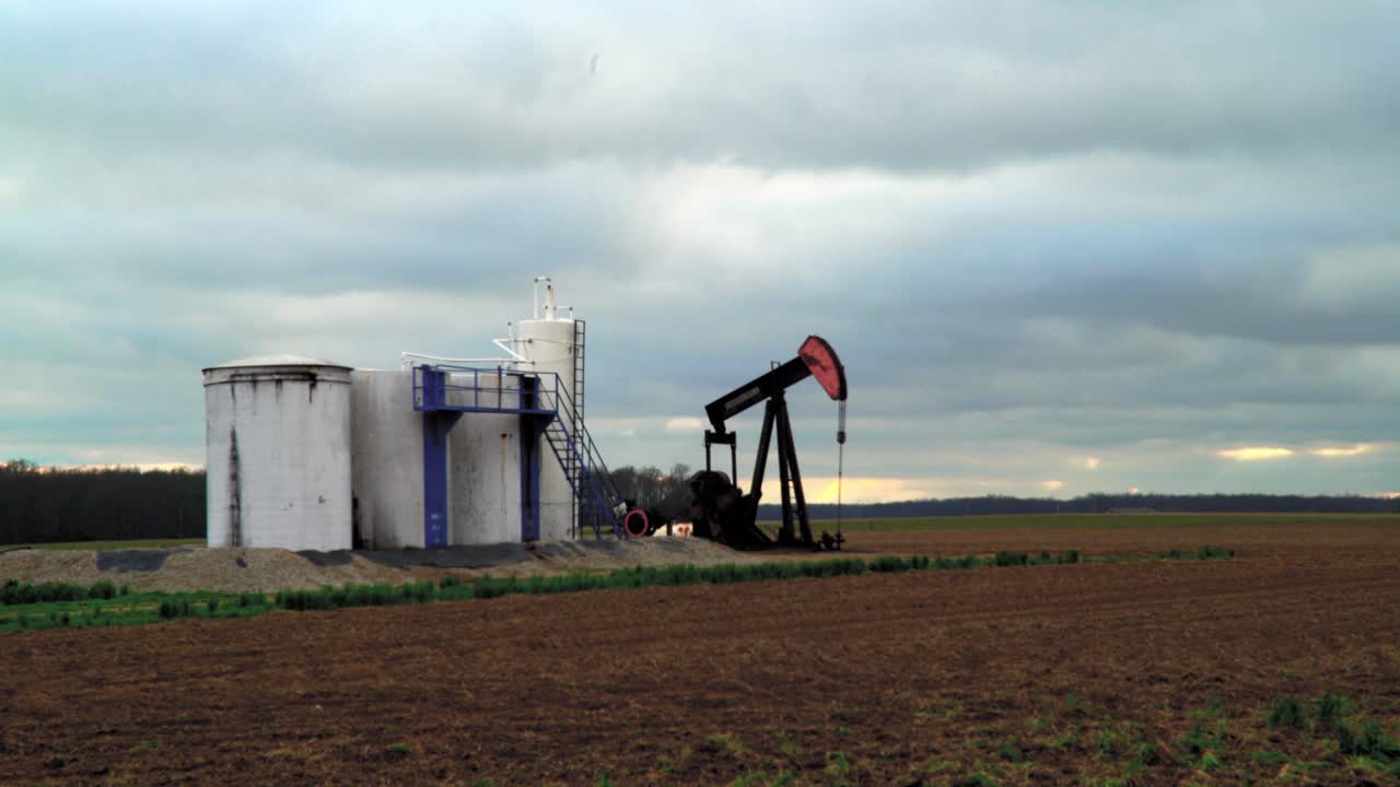 Oil field pump jack slowly pumping oil or natural gas out of the ground with storage facility. 4K UHD Wide Shot.