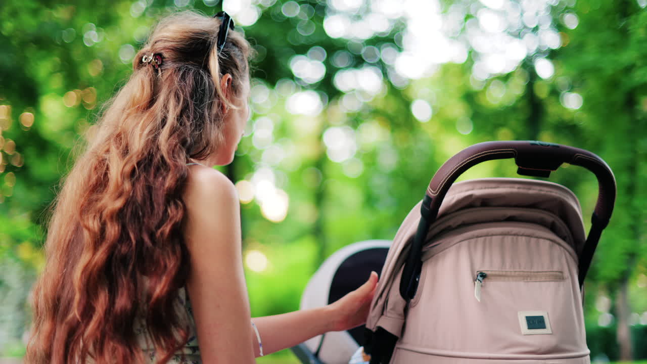 A woman with long curly hair holding a stroller while sitting on a park bench surrounded by greenery