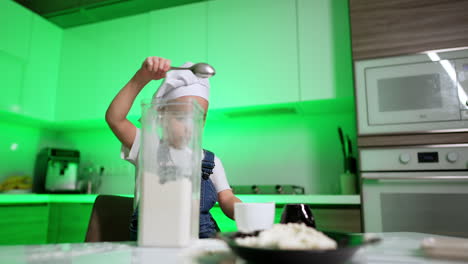 adorable baby boy little chef in the kitchen. focused kid puts flour from container to the cup.