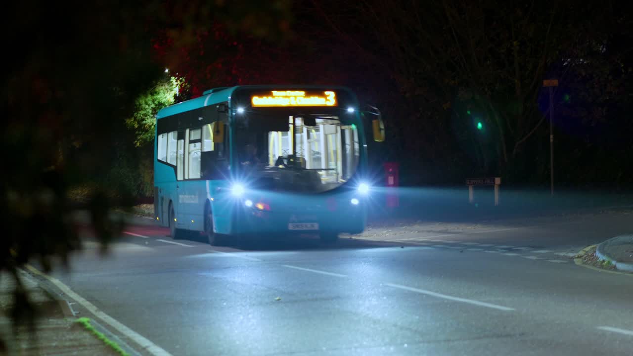 A bus with headlights on drives towards and past the camera in a nighttime setting