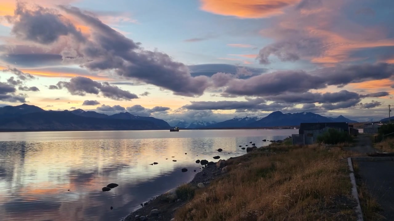Uma noite tranquila da Patagônia se desenrola ao longo do Canal Última Esperança, perto de Puerto Natales, Chile, enquanto nuvens de pôr do sol ardentes projetam reflexos dourados e carmesim sobre as águas calmas do fiorde