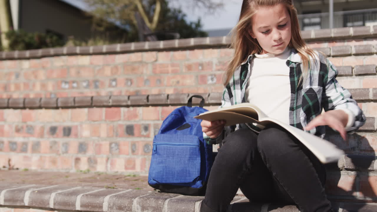 Schoolgirl sitting on steps reading book with blue backpack beside her, copy space