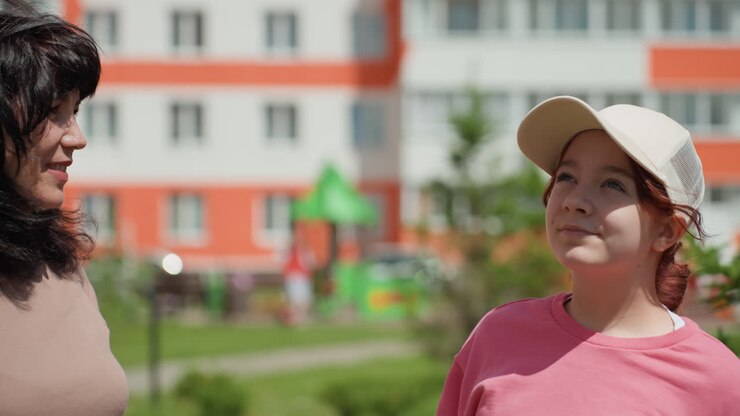 Donna e ragazza che indicano il cielo nel cortile, momento giocoso tra vicini, luce solare intensa, sfondo di un appartamento, area verde e parco giochi visibili, interazione gioiosa e scoperta condivisa