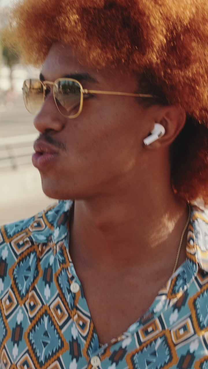 Giovane uomo con capelli ricci e rossi che indossa occhiali da sole e auricolari wireless