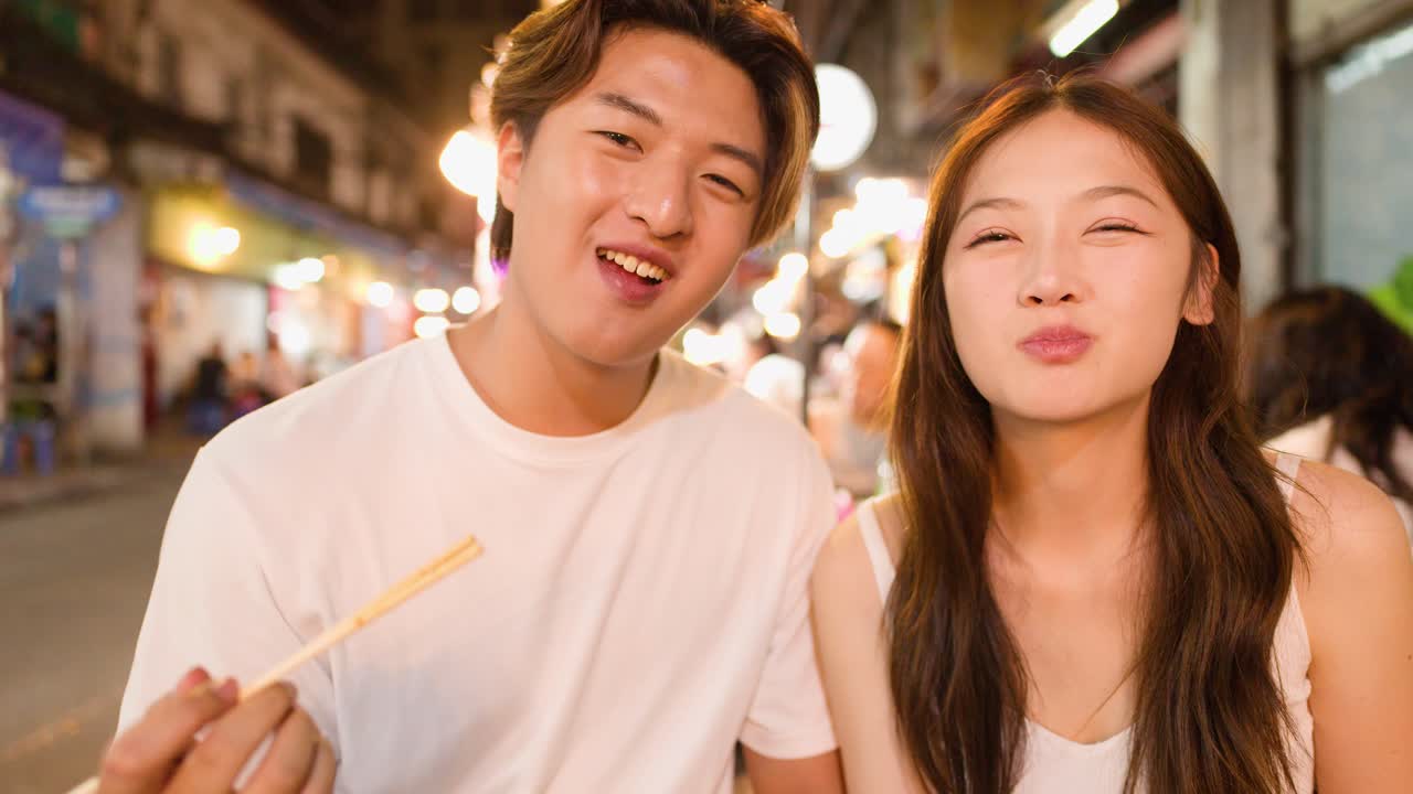 Smiling couple eats noodles with chopsticks at outdoor night market, warm lighting, close-up view