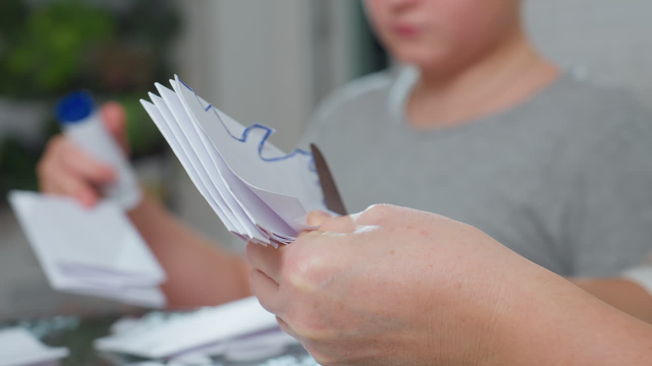 hand view of woman carefully cutting white paper with scissor to create handmade craft while blurred boy in grey shirt seen in background during indoor creative art session at home environment