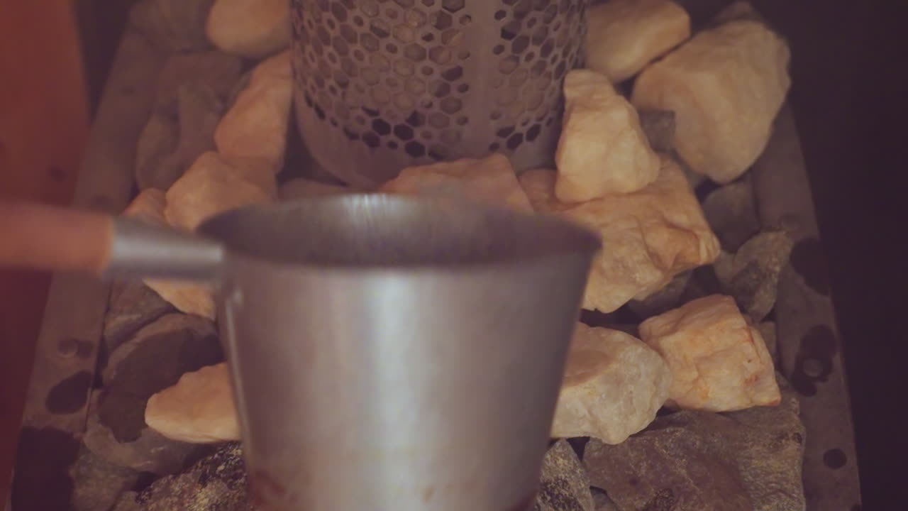 close up of person pouring water onto hot sauna stones with steam rising against wooden wall, metal ladle in focus, ritual of therapeutic heat infusion, calming wellness atmosphere