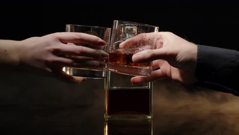 two hands with glasses of cognac whiskey with ice cubes making cheers toast on black background