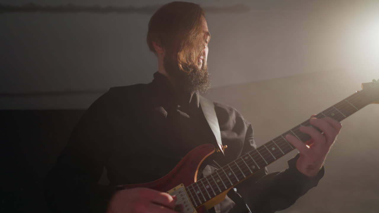 Bearded guitarist in black attire plays a red guitar, gently moving his body in a softly illuminated room