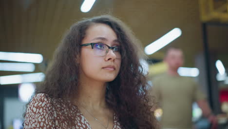 lady with vintage curly hair and glasses focuses intently on item inside her bag in bustling mall environment, soft bokeh lights create a blurred, atmospheric background, with people passing by