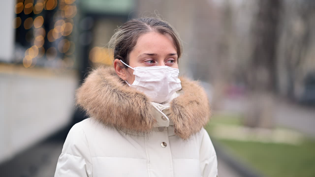 In a winter park, a woman takes a stroll while wearing a face mask. She is dressed warmly with a fur-lined coat, observing her surroundings with a calm demeanor under gray skies